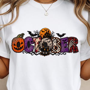 Könnte beinhalten: Ein weißes T-Shirt mit einem grafischen Design des Wortes OCTOBER in großen, stilisierten Buchstaben. Jeder Buchstabe enthält Halloween-Motive wie einen Kürbis, ein Spukhaus, Geister, Fledermäuse, Spinnennetze und eine Skeletthand.