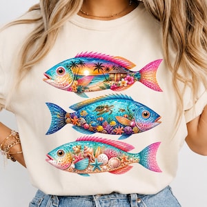 May include: A cream-colored t-shirt featuring a graphic of three stacked fish. The top fish contains a beach sunset scene, the middle fish displays an underwater coral reef with a sea turtle, and the bottom fish shows various seashells and starfish.