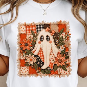 Könnte beinhalten: Ein weißes T-Shirt mit einem quadratischen Patchwork-Design und einer cremefarbenen Geisterfigur in der Mitte. Das Motiv besteht aus orangefarbenen, braunen und schwarzen karierten und floralen Stoffquadraten, gestickten Blumen, grünen Blättern und einer kleinen Schleife mit Knopf.