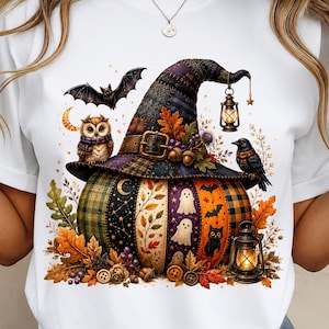 May include: A white t-shirt featuring a graphic of a large pumpkin topped with a patchwork witch hat. The design includes an owl, a bat, a crow, a black cat, ghosts, lanterns, autumn leaves, and buttons, all arranged in a seasonal display.