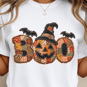 May include: A white t-shirt featuring a graphic of the word BOO in a patchwork style. The letters are filled with orange, brown, and green patterns, including plaid and leopard print. A black witch hat sits on the letter O, and two black bats.