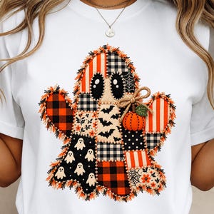 Könnte beinhalten: Ein weißes T-Shirt mit einer großen, zentralen Grafik eines Geistes, der aus verschiedenen Stoffflicken zusammengesetzt ist. Das Geisterdesign enthält Muster aus schwarz-orangem Karomuster, Streifen, Fledermäusen, Spinnennetzen und kleinen weißen Geistern.