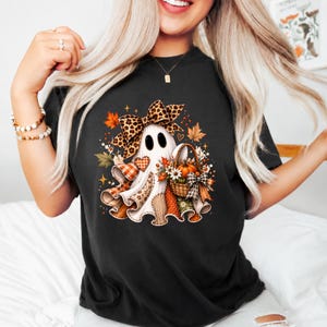 Puede incluir: Una camiseta negra con el gráfico de un fantasma blanco que lleva un lazo grande con estampado de leopardo. El fantasma está rodeado de hojas de otoño, una pequeña cesta llena de calabazas y flores, y varios parches de tela estampada.