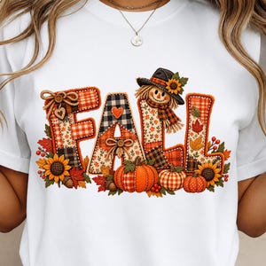 Puede incluir: Una camiseta blanca con un estampado gráfico de la palabra FALL en letras grandes y texturizadas. Las letras están decoradas con patrones de cuadros, vichy y florales, una cara de espantapájaros, calabazas, girasoles y hojas de otoño.
