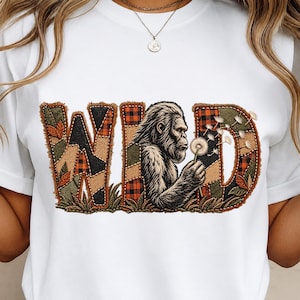 Peut inclure: Un t-shirt blanc avec une grande impression graphique du mot WILD. Les lettres sont remplies d'un motif patchwork de textures de tissu à carreaux orange et noir, vert et marron. Une illustration détaillée d'un Bigfoot tenant un pissenlit.