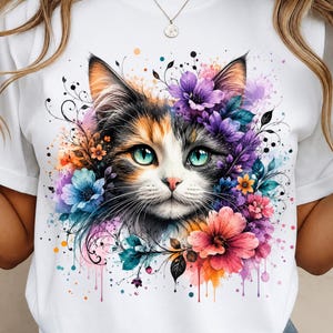 Può includere: Una t-shirt bianca con una stampa grafica colorata di un muso di gatto calico circondato da vivaci fiori in stile acquerello nelle tonalità del viola, rosa, arancione e blu, con dettagli di viticci neri e schizzi di vernice.