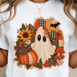 Könnte beinhalten: Ein weißes T-Shirt mit einem zentralen Grafikdesign, das einen cremefarbenen Geist, einen Patchwork-Kürbis, eine Sonnenblume, rote Pilze, eine gelbe Mondsichel mit einer schwarzen Fledermaus sowie verschiedene karierte und florale Stoffmuster zeigt.