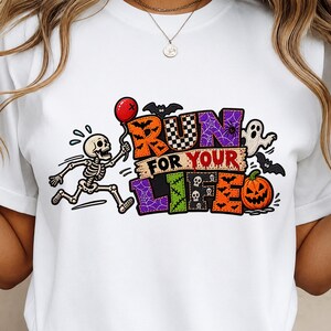 Könnte beinhalten: Ein weißes T-Shirt mit einer bunten Grafik und dem Schriftzug RUN FOR YOUR LIFE. Das Design zeigt ein laufendes Skelett mit einem roten Luftballon, ein Gespenst, Fledermäuse und einen Kürbis in leuchtenden Orange-, Lila- und Grüntönen.