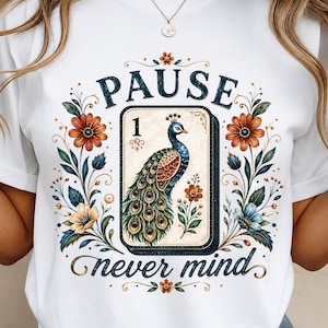 May include: A white t-shirt featuring a central rectangular playing card graphic with the number 1 and a colorful peacock illustration. The design is surrounded by floral motifs and the text PAUSE above and never mind below in dark blue.