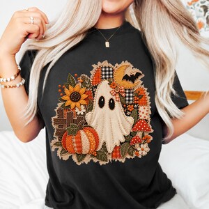 Peut inclure: Un t-shirt noir avec une impression graphique repr&eacute;sentant un fant&ocirc;me, une citrouille, un tournesol, un croissant de lune avec une chauve-souris et des champignons. Le motif int&egrave;gre divers textures &agrave; carreaux orange, blancs et noirs.