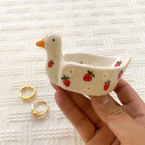 May include: A small, cream-colored ceramic duck-shaped trinket dish featuring a painted pattern of red strawberries and white flowers. The duck has a bright orange beak and a black eye. Two gold-toned metal rings are positioned nearby.