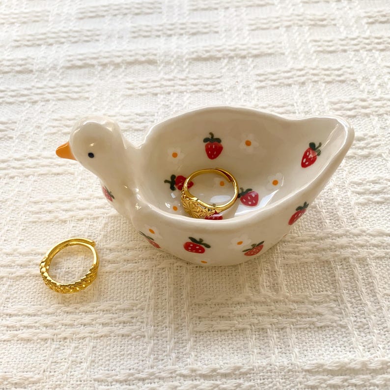 May include: A cream-colored ceramic duck-shaped trinket dish featuring a hand-painted pattern of small red strawberries and white flowers. One gold-toned textured ring rests inside the dish, while a second matching gold ring sits beside it.