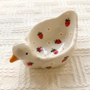 May include: A small, cream-colored ceramic dish shaped like a duck with an orange beak and black eyes. The interior and exterior are decorated with a repeating pattern of small red strawberries and tiny white flowers with yellow centers.