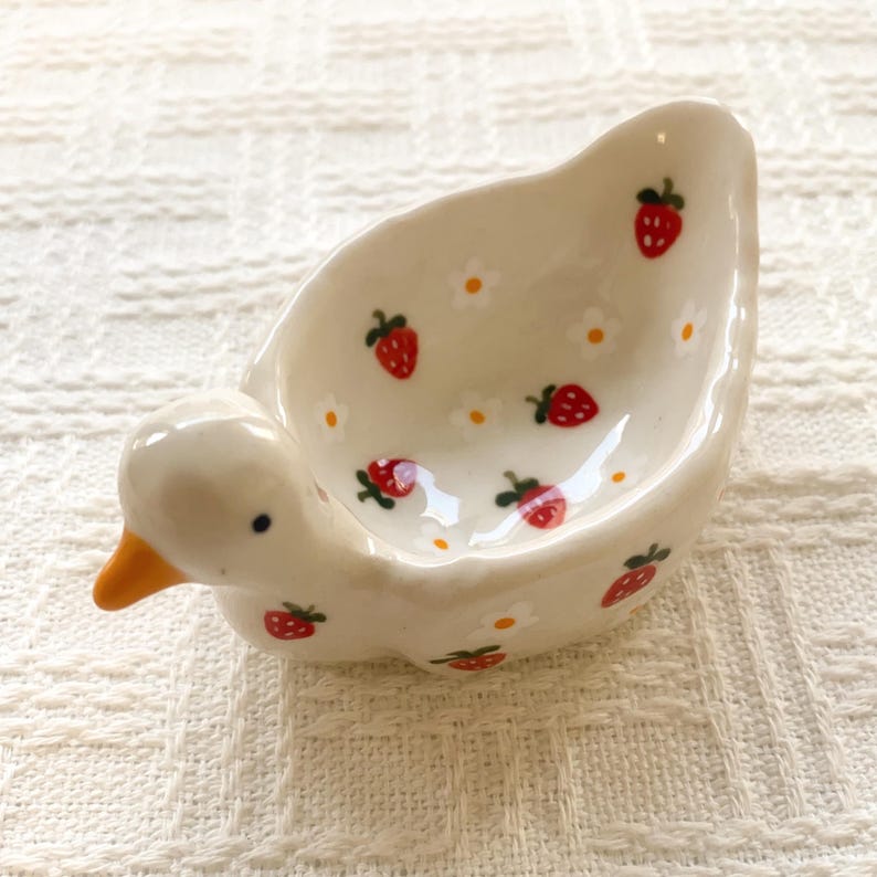 May include: A small, cream-colored ceramic dish shaped like a duck with an orange beak and black eyes. The interior and exterior are decorated with a repeating pattern of small red strawberries and tiny white flowers with yellow centers.
