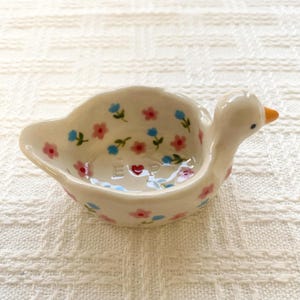 Peut inclure: Un petit vide-poche en céramique couleur crème en forme de canard. L'intérieur présente un motif peint à la main de minuscules fleurs roses et bleues avec des tiges vertes. Les lettres E et P sont gravées au centre, séparées par un cœur rouge.