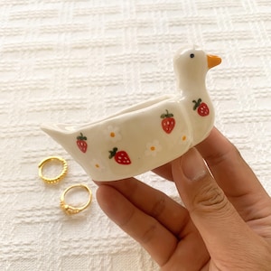 May include: A cream-colored ceramic duck-shaped dish featuring a hand-painted pattern of small red strawberries and white flowers. The duck has a bright orange beak and a black eye. The dish is designed with an open, hollow center.