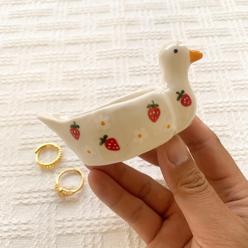 May include: A cream-colored ceramic duck-shaped dish featuring a hand-painted pattern of small red strawberries and white flowers. The duck has a bright orange beak and a black eye. The dish is designed with an open, hollow center.