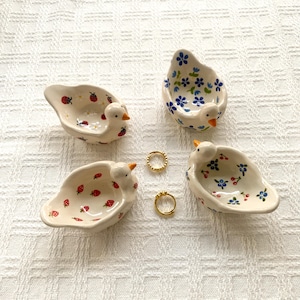 May include: Four small, white, duck-shaped ceramic dishes with hand-painted patterns including red ladybugs, blue flowers, red strawberries, and blue berries with green leaves. Two small gold-toned metal rings are placed in the center.