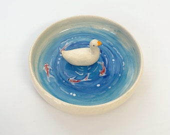 May include: A round ceramic dish featuring a hand-painted blue water design with white ripples. A small, three-dimensional white duck figurine with an orange beak is attached to the center, surrounded by painted orange and white koi fish.