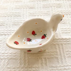 May include: A small, off-white ceramic dish shaped like a bird, featuring a hand-painted pattern of red strawberries and white flowers with yellow centers. The bird-shaped bowl has a smooth, glossy finish and a small orange beak detail.