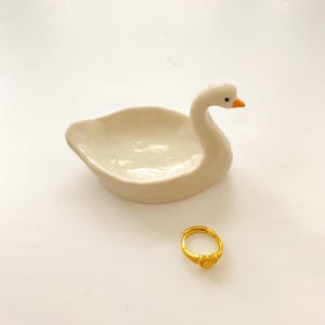 May include: A cream-coloured ceramic swan-shaped trinket dish with a smooth, glossy finish and an orange beak, positioned next to a small, circular gold-toned ring featuring a textured, heart-shaped design on the front of the band.