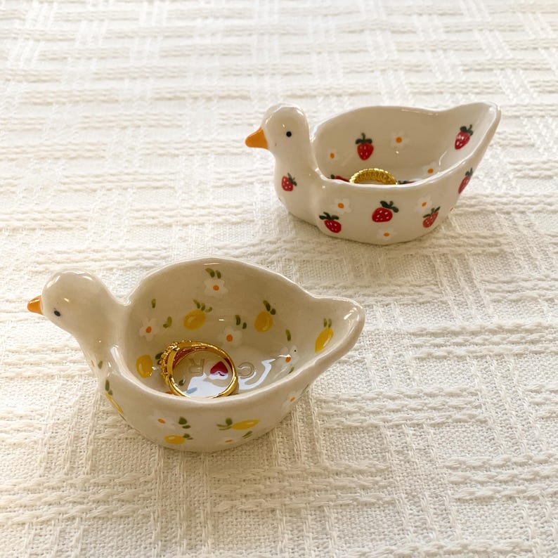 May include: Two ceramic duck-shaped trinket dishes. One dish features a white base with red strawberry and white flower patterns, containing a gold ring. The other dish has a white base with yellow lemon and white flower patterns, containing a gold ring.