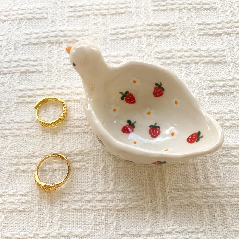 May include: A cream-colored ceramic bird-shaped trinket dish featuring a hand-painted pattern of small red strawberries and white flowers with yellow centers, accompanied by two gold-toned textured hoop rings placed to the left of the dish.