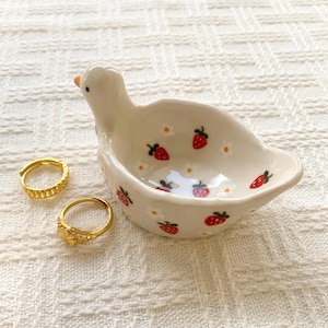 May include: A white ceramic trinket dish shaped like a duck with a painted pattern of red strawberries and small white flowers, accompanied by two gold-toned metal rings with textured surfaces resting beside the dish.