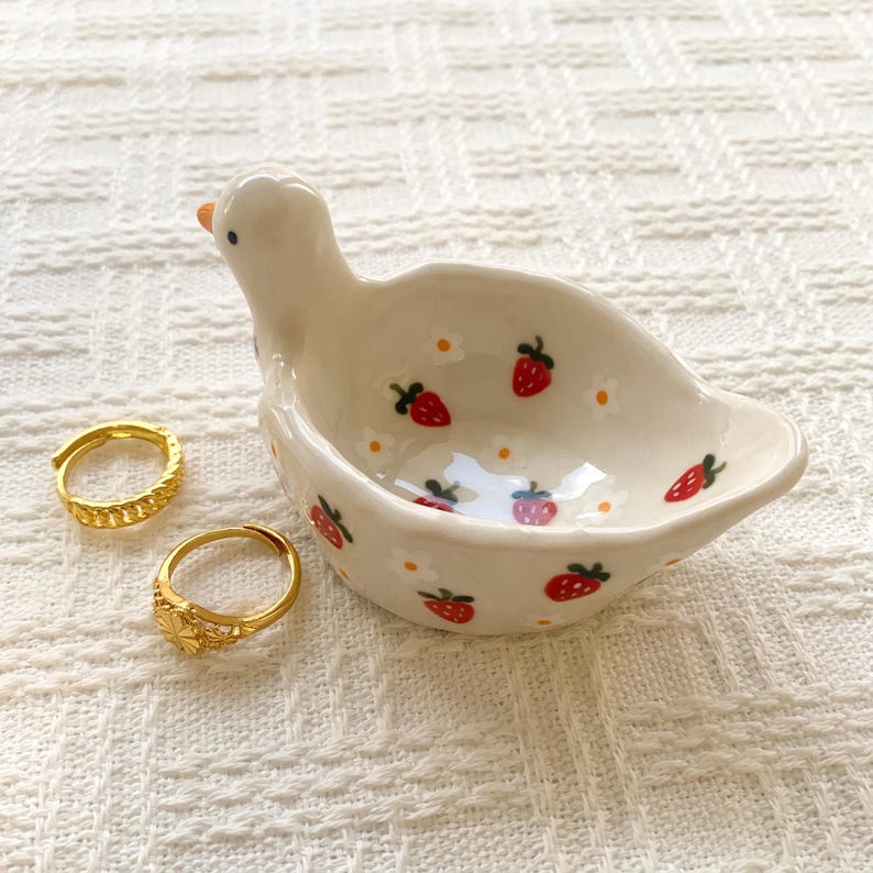 May include: A white ceramic trinket dish shaped like a duck with a painted pattern of red strawberries and small white flowers, accompanied by two gold-toned metal rings with textured surfaces resting beside the dish.