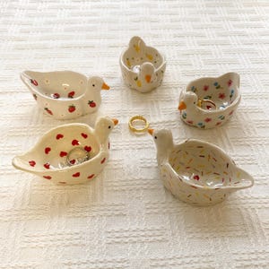 May include: Five small ceramic duck-shaped trinket dishes with various patterns including red strawberries, yellow lemons, colorful flowers, red hearts, and multicolored sprinkles, with two gold-toned metal rings placed inside.