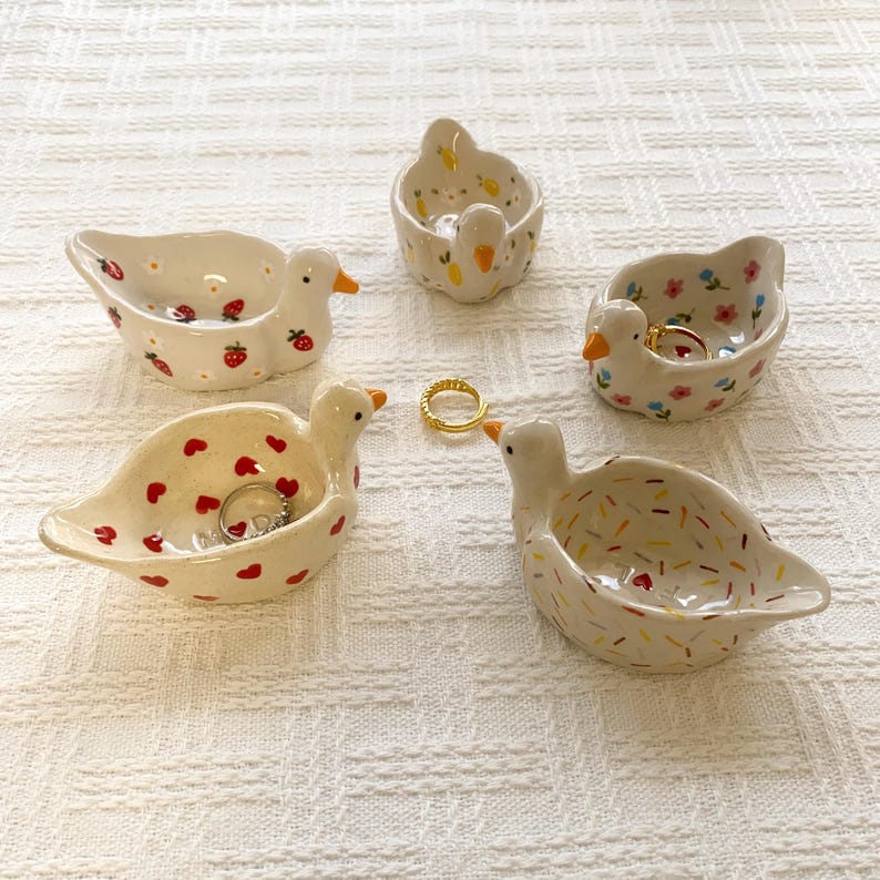 May include: Five small ceramic duck-shaped trinket dishes with various patterns including red strawberries, yellow lemons, colorful flowers, red hearts, and multicolored sprinkles, with two gold-toned metal rings placed inside.