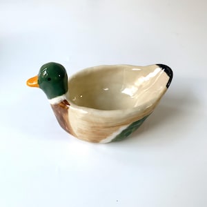 May include: A ceramic bowl shaped like a mallard duck. The duck head features a dark green neck, a bright orange beak, and a small black eye. The body of the bowl is cream-colored with brown and green painted details, and a black-tipped tail.