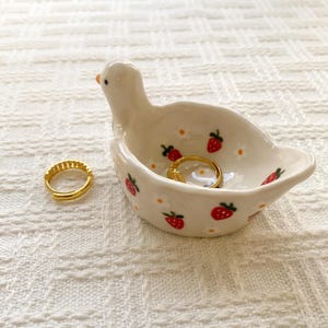 May include: A white ceramic trinket dish shaped like a duck, decorated with small red strawberries and white flowers. The dish contains one gold hoop earring, and a second matching gold hoop earring sits on the surface next to the duck figure.