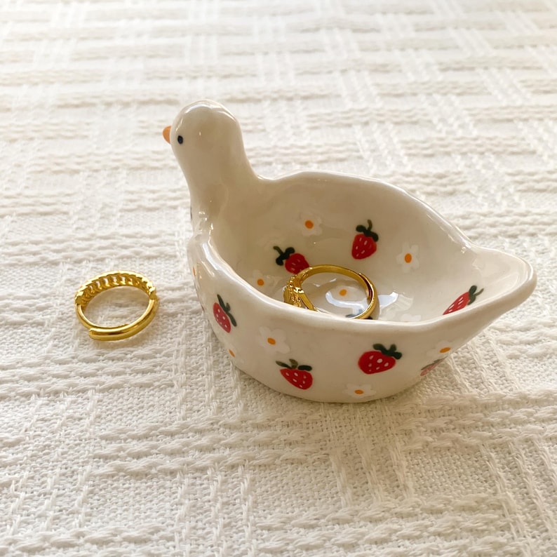 May include: A white ceramic trinket dish shaped like a duck, decorated with small red strawberries and white flowers. The dish contains one gold hoop earring, and a second matching gold hoop earring sits on the surface next to the duck figure.