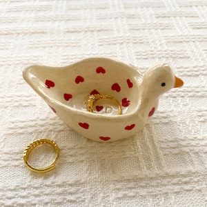 May include: A cream-colored ceramic duck-shaped trinket dish featuring small red heart patterns across its surface. The dish contains one gold hoop earring, and a second matching gold hoop earring is positioned separately on the surface.