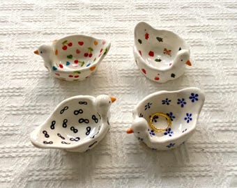 May include: Four small, white ceramic bird-shaped dishes, each featuring a unique hand-painted pattern: red cherries, colorful abstract shapes, black infinity symbols, and blue flowers. One dish contains a small gold-toned ring.