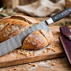 May include: A Damascus steel bread knife with a dark wood handle is partially slicing through a round loaf of crusty sourdough bread. A dark purple leather sheath with decorative stitching and a metal snap closure rests beside the bread.