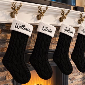 May include: Four black cable-knit Christmas stockings with white ribbed cuffs, each personalized with black embroidered names William, Olivia, Julian, and Amelia, hanging from gold-colored metal deer head hooks on a white fireplace mantel.