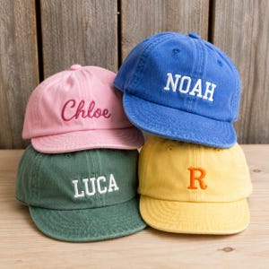 Op de afbeelding: Vier baseballpetten in roze, blauw, groen en geel. De roze pet heeft de geborduurde naam Chloe, de blauwe pet toont de naam NOAH, de groene pet laat de naam LUCA zien en de gele pet bevat de enkele letter R.