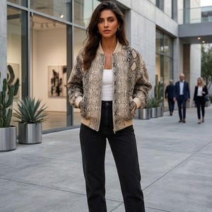 May include: A beige and brown snakeskin print bomber jacket with ribbed cuffs and hem, worn over a plain white tank top and high-waisted black denim jeans. The jacket features a front zipper closure and a classic collarless neckline design.