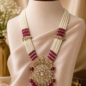 May include: A multi-strand necklace featuring rows of small white beads interspersed with sections of deep red beads. The necklace includes a large, ornate gold-toned pendant encrusted with clear stones and accented with dangling red teardrop beads.