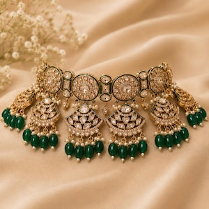 May include: A gold-toned choker necklace featuring circular and fan-shaped motifs embedded with clear crystals. The piece is accented with dark green teardrop-shaped beads and small white pearls hanging from the lower edges of the metal segments.