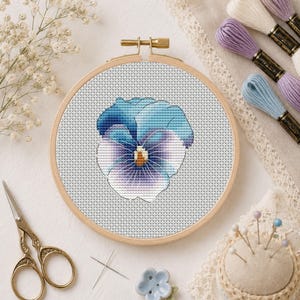 May include: A round wooden embroidery hoop containing a cross-stitch pattern of a blue and purple pansy flower. The design features a yellow centre on a light grey fabric background. The hoop is surrounded by embroidery floss, scissors, and a pin cushion.