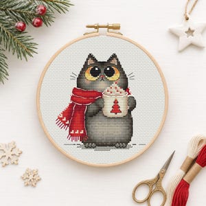 Op de afbeelding: Een kruissteek borduurproject in een houten borduurring met een ronde, grijze kat met grote gele ogen die een rode sjaal met witte hartjespatronen draagt. De kat houdt een witte mok vast die is versierd met een rode kerstboom.