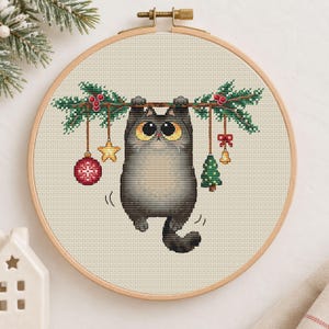 May include: A cross-stitch embroidery in a round wooden hoop featuring a gray cat with large yellow eyes hanging from a green pine branch. The branch is decorated with a red patterned ornament, a yellow star, a small tree, and a bell.