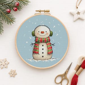 May include: A cross-stitch embroidery in a wooden hoop featuring a snowman with a carrot nose, green earmuffs, and a red and green striped scarf against a light blue background with white dots representing falling snow.