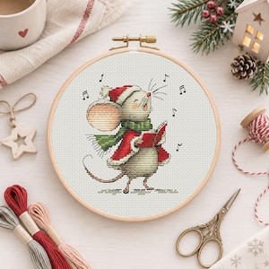 May include: A circular wooden embroidery hoop containing a cross-stitch pattern of a mouse wearing a red Santa hat and a green scarf, singing while holding a red book. Musical notes are stitched around the mouse on white fabric.