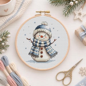 May include: A cross-stitch embroidery project in a round wooden hoop featuring a snowman wearing a blue and white striped knit hat and matching scarf. The snowman has a small orange carrot nose, two dark eyes, and three brown buttons.