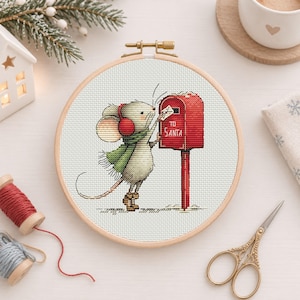 May include: A circular wooden embroidery hoop containing a cross-stitch pattern of a light gray mouse wearing red earmuffs, a green scarf, and brown boots. The mouse is placing a letter into a red mailbox labeled TO SANTA.
