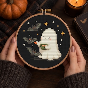 May include: A circular wooden embroidery hoop containing a cross-stitch design on black fabric. The design features a white ghost holding a green bowl with orange contents, surrounded by three grey bats and several small yellow star-like shapes.